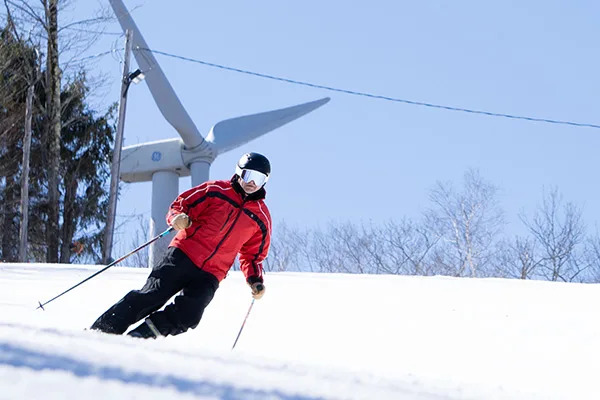 A skier descends a Jiminy Peak trail with the Zephyr wind turbine standing on the summit ridge above